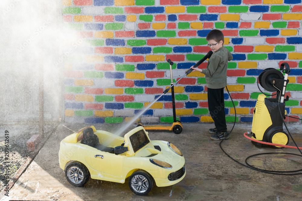 Obraz premium Happy child washes yellow toy car from high pressure washer in front of a multi-colored brick wall. Fun during the summer holidays. The concept of family business.