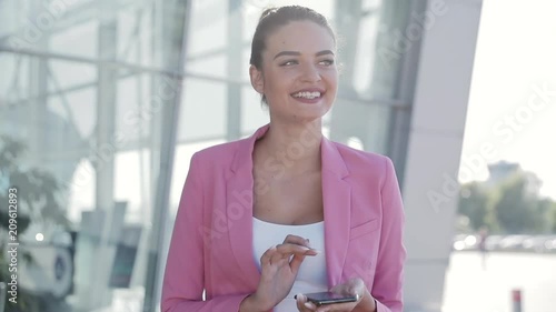 Female portrait of business worker. Using her Modern Smartphone, wearing Classical Jacket. Looks Successful and Confident. Laughing sincerly. Office building in the background. photo