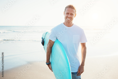 Portrait of happy surfer in hawaiian t-shirt walking with surf board on the beach at sunset photo