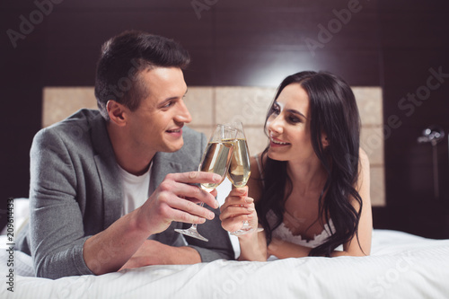 For our young family. Portrait of excited bride and groom are celebrating their wedding day with festive beverage. They are clinking glasses and smiling while lying on bed 
