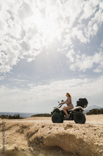 Young woman driving rental quad bike on seaside road in Milos island towards Firiplaka beach, Greece