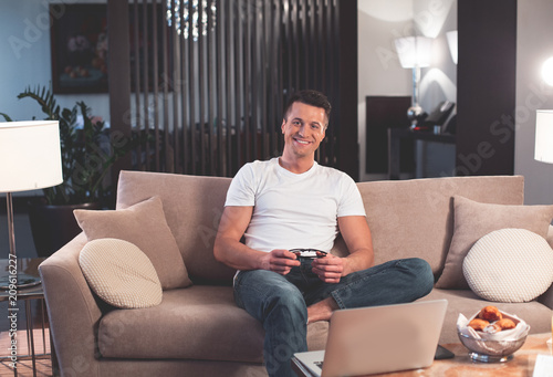 I am ready to work at home. Portrait of cheerful male freelancer is sitting on sofa near his laptop and holding eyeglasses. He is looking at camera with joy and laughing 