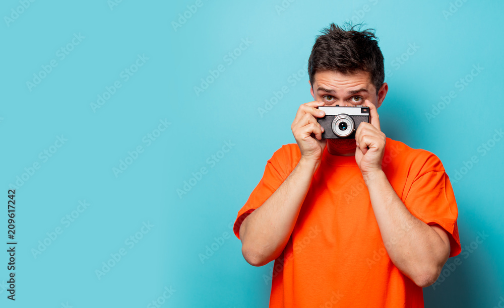 Young handsome man in orange t-shirt with vintage camera. Studio image on blue background Young handsome man in orange t-shirt with vintage camera. Studio image on blue background