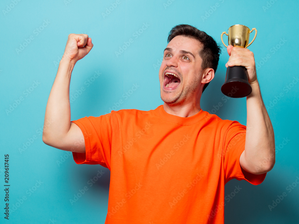 Young handsome man in orange t-shirt with golden cup. Studio image on blue background Young handsome man in orange t-shirt with golden cup. Studio image on blue background