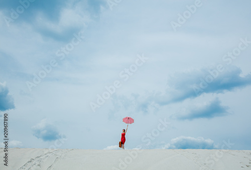 Young woman in red dress with umbrella and suitcase on the beach. Travel concept image on sand