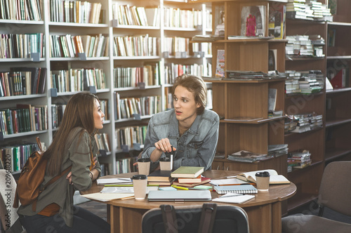 Serious male and orderly female talking with each other while sitting at table in library. They studying with books and cups of coffee