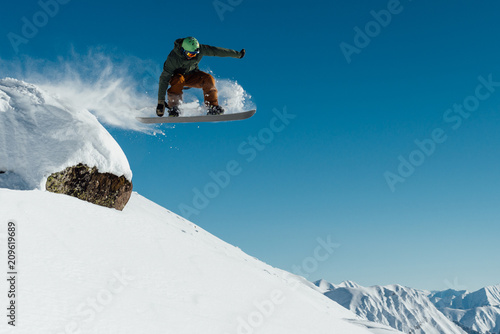   snowboarder in the outfit drops off the ledge of the stone onto the fresh snow creating a spray of snow