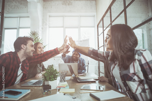 Partnership. Low angle of positive young creative team are sitting at table in office and demonstrating unity and collaboration. They are giving five with smile while enjoying success. Selective focus photo