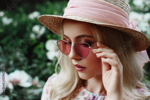 Outdoor close up portrait of beautiful young woman wearing pink sunglasses, straw hat posing in the blooming garden. Model looking down. Copy, empty space for text