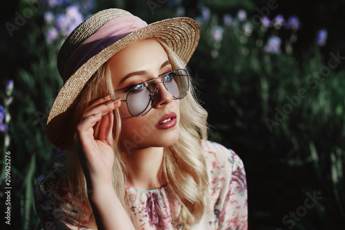 Outdoor close up portrait of beautiful young woman wearing blue sunglasses, straw hat posing in the blooming garden. Model looking aside. Copy, empty space for text