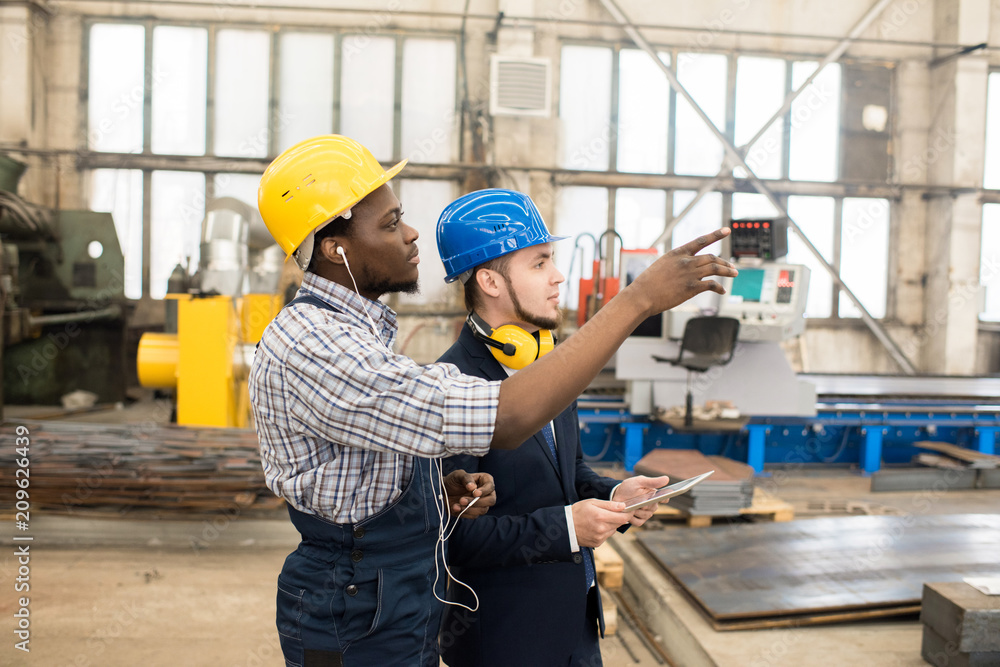 Serious confident African-American factory worker in earphones wearing hardhat showing equipment to inspector in formal suit wearing soundproof headphones using tablet for notes in workshop. Serious confident African-American factory worker in earphones wearing hardhat showing equipment to inspector in formal suit wearing soundproof headphones using tablet for notes in workshop.