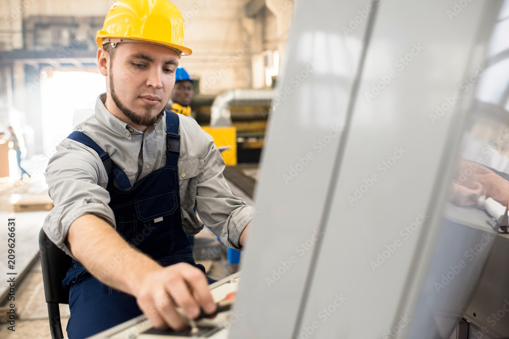 Serious concentrated bearded machine operator in yellow hardhat using control panel while managing conveyor belt at factory workshop Serious concentrated bearded machine operator in yellow hardhat using control panel while managing conveyor belt at factory workshop