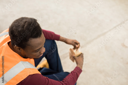 Directly above of pensive calm African-American construction worker in orange vest sitting on ground and eating from box during lunch break