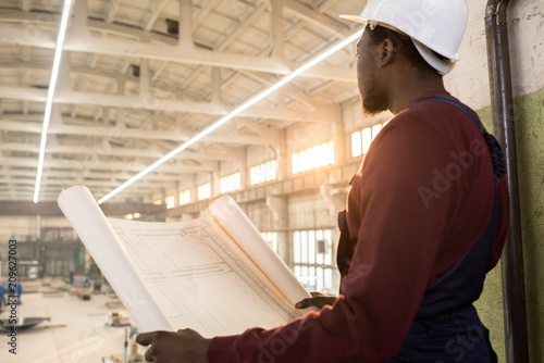 Rear view of pensive purposeful young African-American construction manager in hardhat holding blueprint with building plan and looking around at construction site