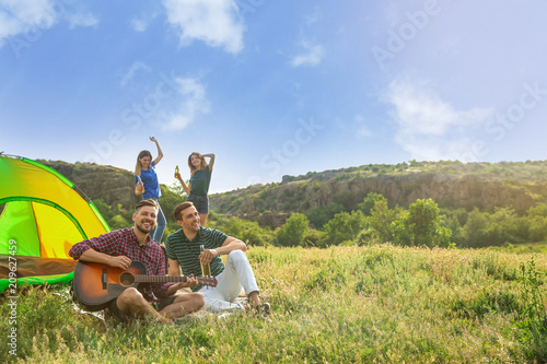 Group of young people resting with beer and guitar near camping tent in wilderness