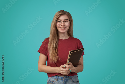Young smiling lady student in eyeglasses and red shirt standing with folder in hands and happily looking aside on over colorful background © EverGrump