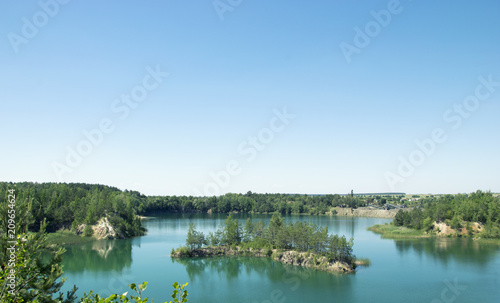 An island in the shape of an arc in the middle of a large lake near the rocks, Blue clear sky An island with trees and forestland, Reflection in the water © Valentine
