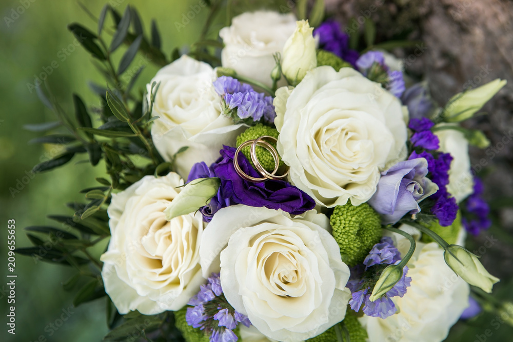 White roses with blue and lavender flowers bouquet. Wedding ring macro photography. Beautiful bridal bouquet with white roses and golden rings