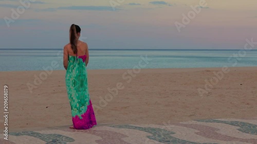 Ragazza guarda mare in estate. Vestito lungo colorato. Cielo al tramonto blue e arancione. Concettuale eleganza, libertà e gioventù