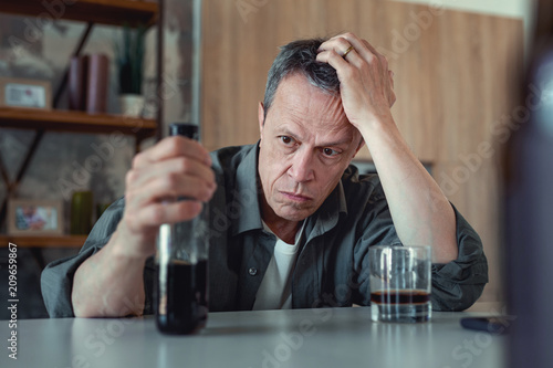 Bottle of cognac. Dark-eyed mature man looking at the bottle of cognac after stressful events sitting in the kitchen