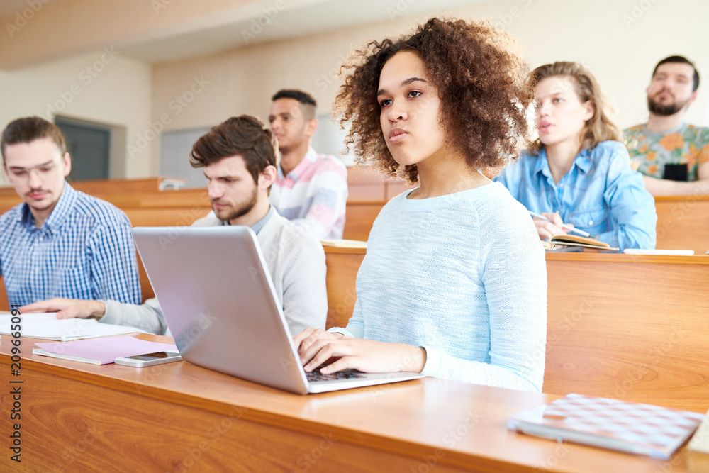 Pensive concentrated young multiethnic students sitting at university desk and attentively listening to lecturer, serious African-American girl with Afro hairstyle typing on laptop Pensive concentrated young multiethnic students sitting at university desk and attentively listening to lecturer, serious African-American girl with Afro hairstyle typing on laptop