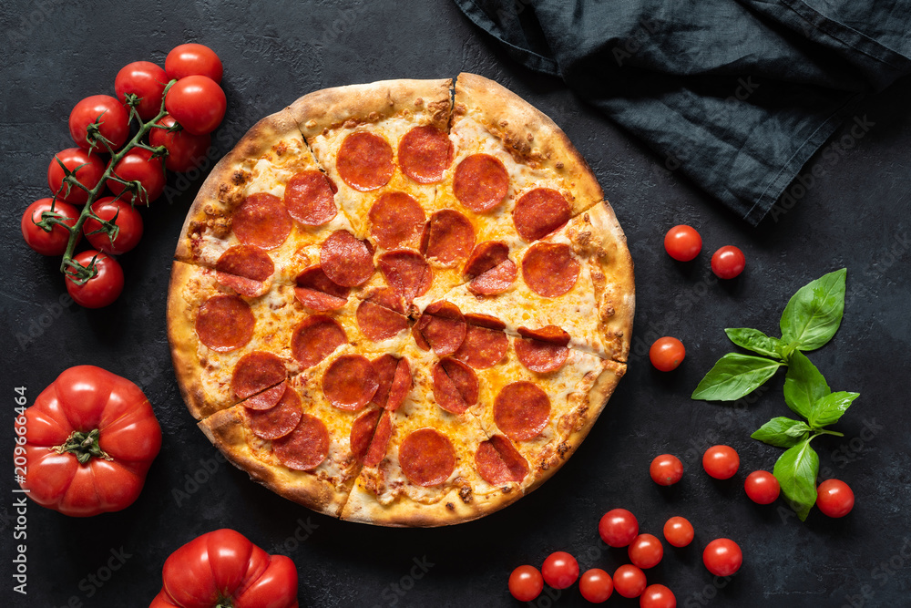 Hot pepperoni pizza on black stone background. Sliced tasty pizza with salami, cheese and tomatoes on dark table, top view