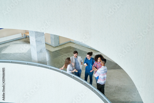 Top view of confident interracial student friends in casual clothing standing at railing in university hall and chatting while waiting to start class photo