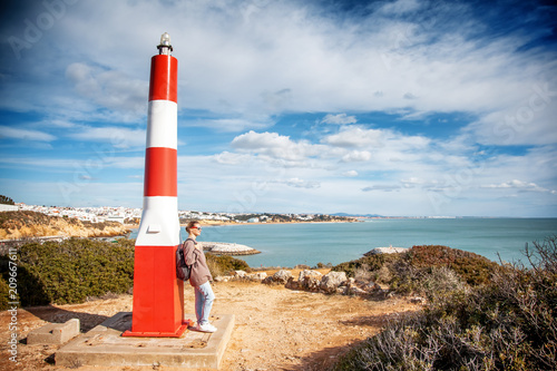 A girl traveler stands by the lighthouse atop a hill and looks at the ocean, the Algarve, Portugal, a popular destination for travel in Europe