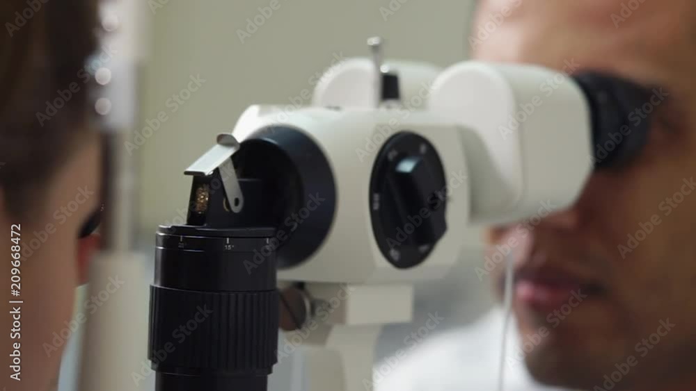 Faces of male eye doctor and patient woman on sides of microscope apparatus. Examining of eyes health by traditional biomicroscopy method Faces of male eye doctor and patient woman on sides of microscope apparatus. Examining of eyes health by traditional biomicroscopy method