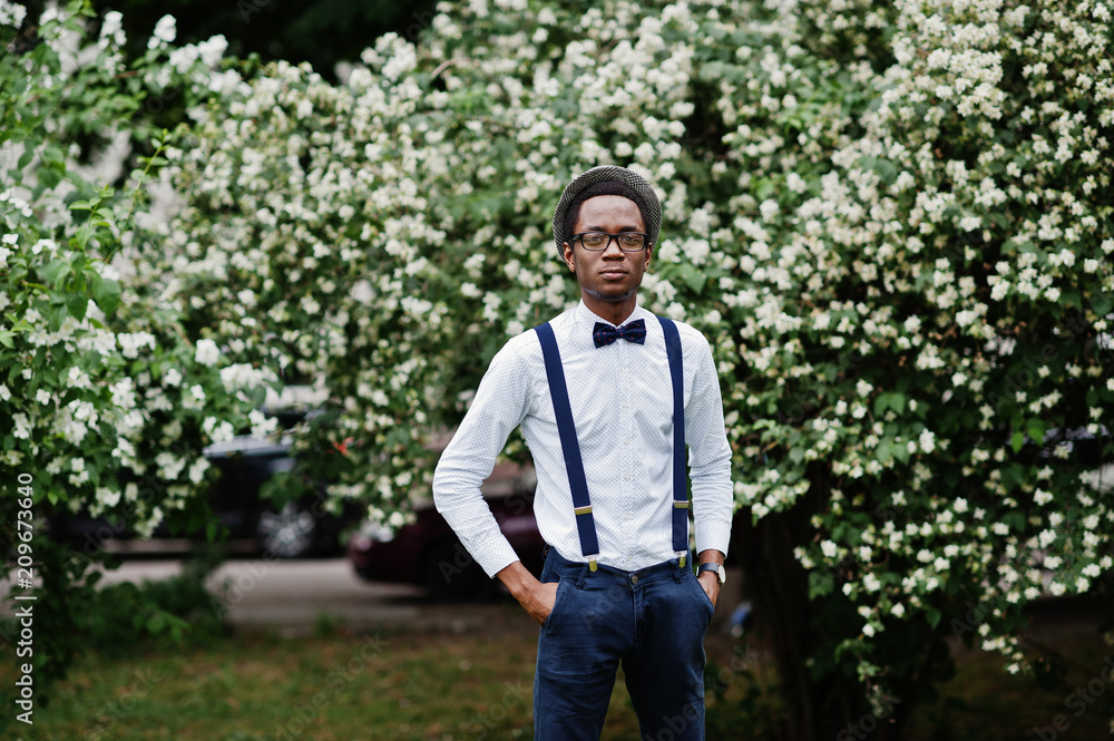 Stylish african american business man at pants with suspender and shirt with bow tie, hat and glasses posed outdoor.