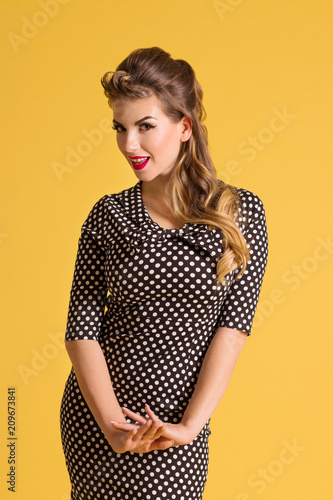Pretty young woman in dress with hairdo stands in yellow studio, pin up style