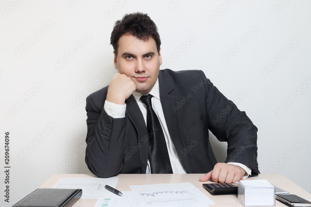 Smiling businessman in suit props his chin at table with notebook, paper, pen, calculator Smiling businessman in suit props his chin at table with notebook, paper, pen, calculator