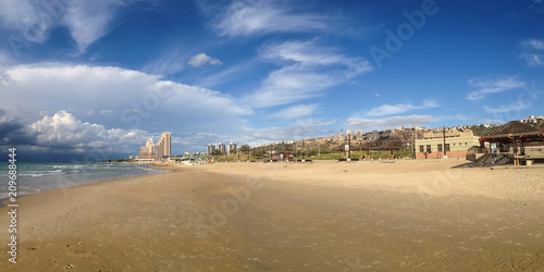 A panoramic view of a lonely romantic sand beach with a beautiful Mediterranean sea and high buildings on the horizon under the blue sky. The photo was shot in a public beach of Haifa, Israel. 