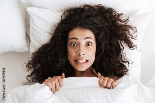 Photo from above of amusing uptight woman 20s with dark curly hair grimacing at camera, lying in bed under white blanket