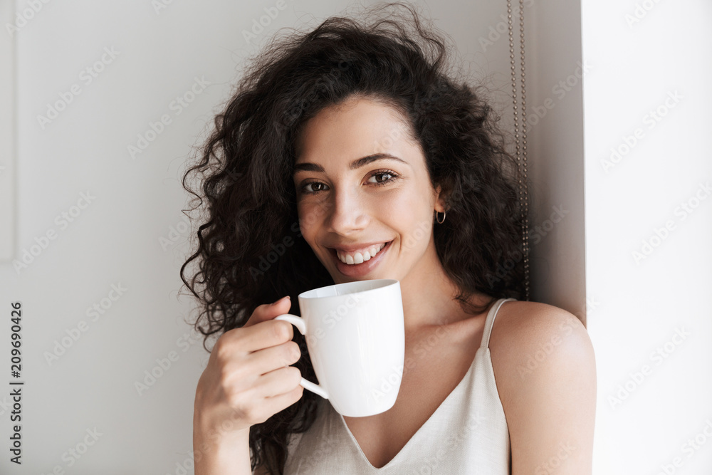 Portrait closeup of gorgeous happy woman with long curly hair smiling at camera, and drinking tea from cup in morning at hotel apartment Portrait closeup of gorgeous happy woman with long curly hair smiling at camera, and drinking tea from cup in morning at hotel apartment