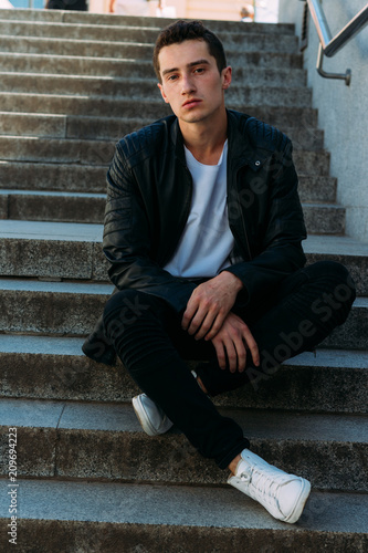 Sexy man posing sits on the steps near railing. Handsome young man in stylish black clothes and white shoes posing near a railing © Skripnik Olga