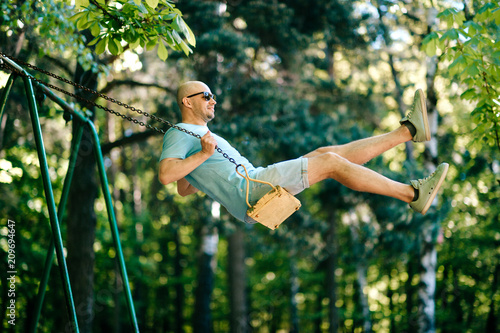 Adult stylish man in glasses riding on swing in city park on playground for children in summer. Happy guy remember childhood. Male person on attraction enjoying up and down motion. Odd boy outdoor.