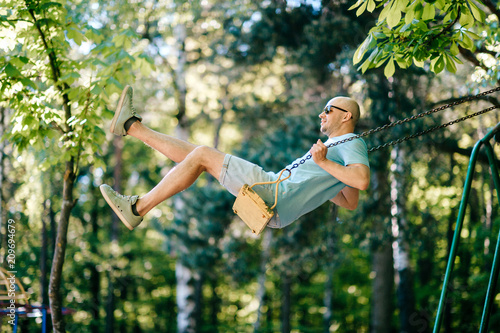 Adult stylish man in glasses riding on swing in city park on playground for children in summer.  Happy guy remember childhood.  Male person on attraction enjoying up and down motion.  Odd boy outdoor.