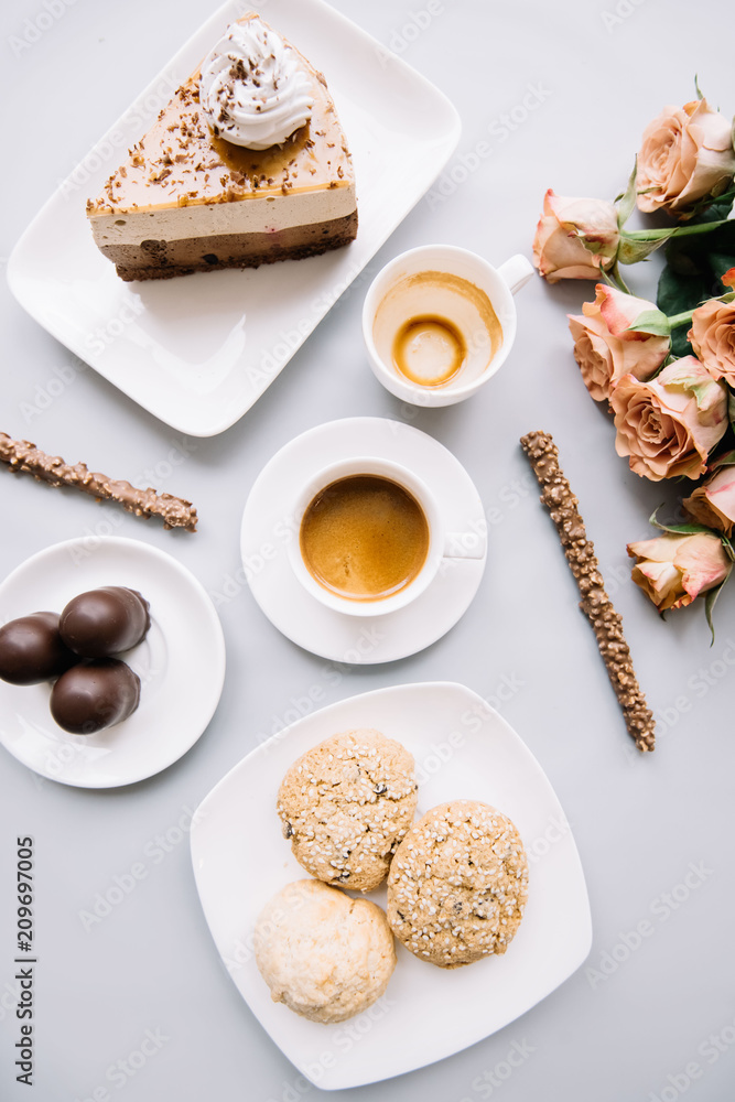 Delicious fresh morning espresso coffee, three cookies, a piece of cake, chocolate sticks, three chocolates and a bouquet of roses on the pastel grey background, top view, flat lay