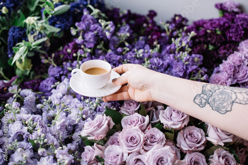 VORONEZH RUSSIA-03.05.2018  Woman with a tattooed hand holding a cup of fresh morning espresso coffee in a white cup with purple colored flowers on the background  roses  carnations  mattiola  tulips 