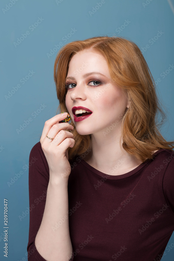 Bright beautiful sexy redhead girl eats with closed eyes colorful candy close-up, blue background, makeup Bright beautiful sexy redhead girl eats with closed eyes colorful candy close-up, blue background, makeup