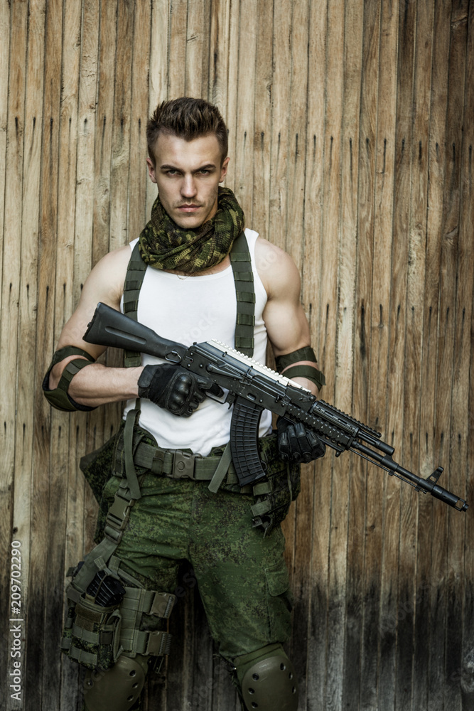 Fototapeta premium Young male military soldier with a rifle posing against wooden background