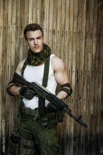 Young male military soldier with a rifle posing against wooden background