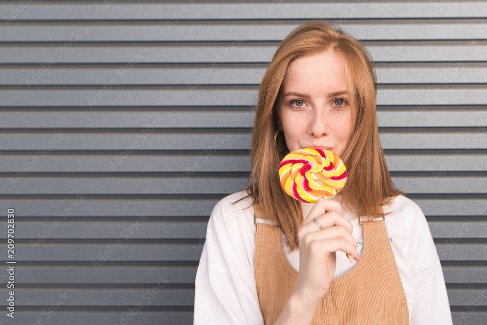 Redheaded girl with freckles eating sweet lollipop and looking at the camera on a background of gray walls. Portrait of a girl with a lollipop on a gray background