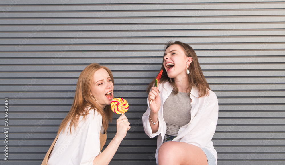 Two playful pretty girls with lollipops in their hands against a gray wall background. Attractive smiling young women with a bow tie in their hands on a gray background Two playful pretty girls with lollipops in their hands against a gray wall background. Attractive smiling young women with a bow tie in their hands on a gray background