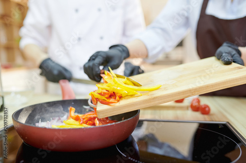 Closeup of modern professional chef putting ingredients into frying pan with gloved hands in modern restaurant kitchen, copy space