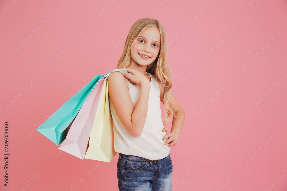 Blond caucasian girl 8-10 in casual clothing smiling at camera and holding colorful shopping bags with purchases, isolated over pink background
