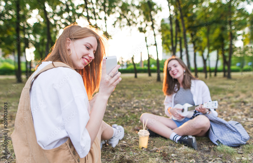 Two pretty happy friends are sitting in the park on the background of the sunset and have a rest. Girl with a smartphone on the background of a girl with ukulele. Leisure with a girlfriend.