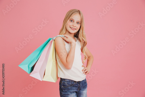 Blond caucasian girl 8-10 in casual clothing smiling at camera and holding colorful shopping bags with purchases, isolated over pink background