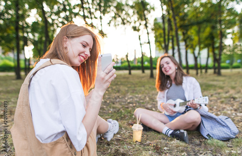 Two pretty happy friends are sitting in the park on the background of the sunset and have a rest. Girl with a smartphone on the background of a girl with ukulele. Leisure with a girlfriend.